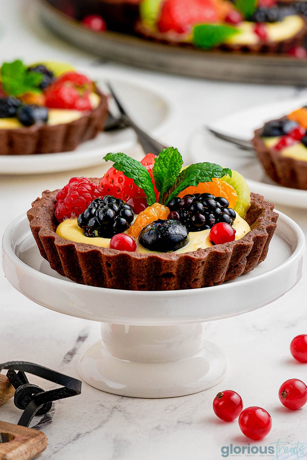 Fruit tart with chocolate shell sitting on small white pedestal topped with various fruit and a shiny passion fruit glaze.