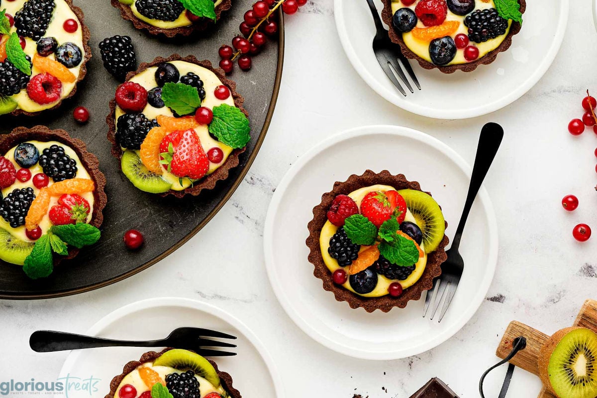 Wide overhead shot of a handful of fruit tartlets with chocolate crusts served on round white plates and more still on a dark round serving tray.