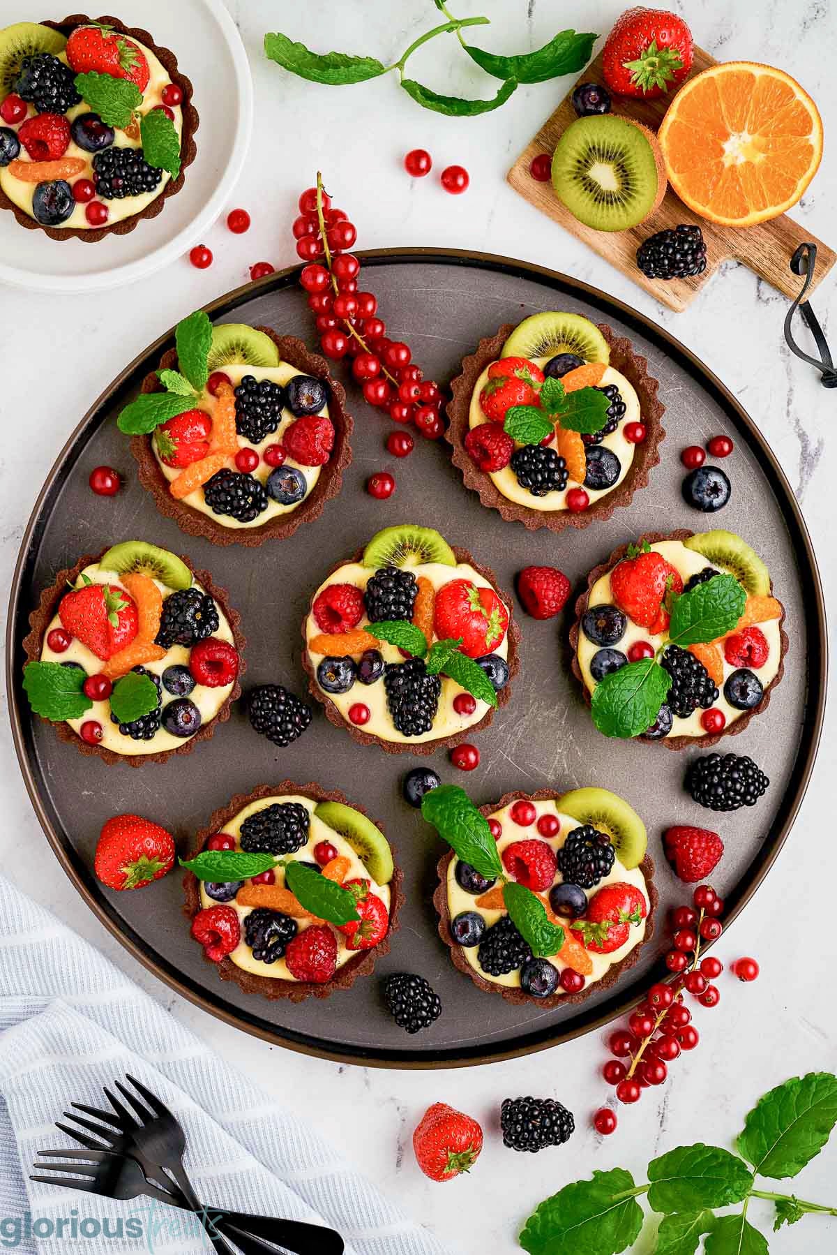 Overhead shot of 7 fruit tarts with chocolate crusts on a round serving platter garnished with mint leaves and more fresh fruit.
